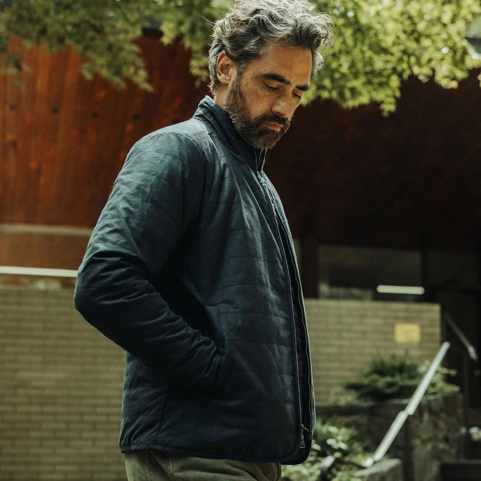 A man with a salt and pepper hair and beard wearing Flint and Tinder's York Quilted Waxed Jacket in Navy. He is shown slight in profile, looking down at his feet with his hands tucked inside the jacket's pockets. A white brick wall, silver stairwell rail and some tree branches appear in the background.