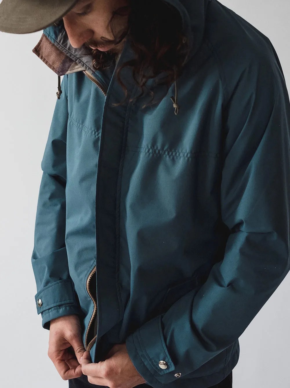 A man wearing a Freeman rain jacket in a bluish green color. The man is shown working to connect the bottom zipper and zip up the jacket. He is wearing a baseball hat and looking down at his fingers working on the zipper.