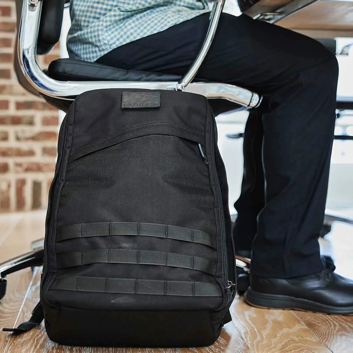 A black GoRuck GR1 shown leaning against a metal swiveling desk chair. A person is sitting in the chair with their legs flat on the floor. 