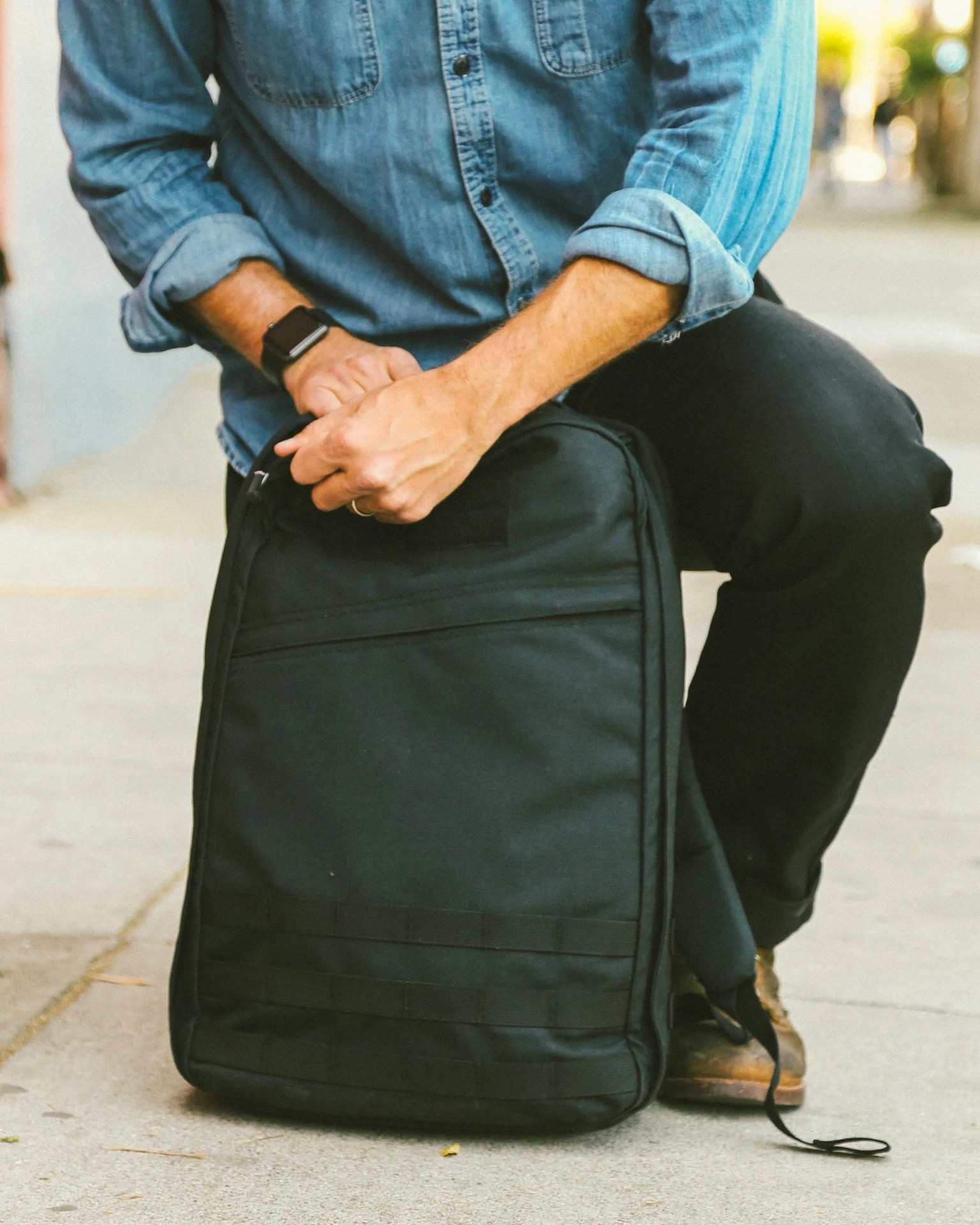 A man shown kneeling in front of a GoRuck GR1 with his hands on the top of the bag looking to open it. The Man's torso is shown wearing a denim shirt with black pants. 