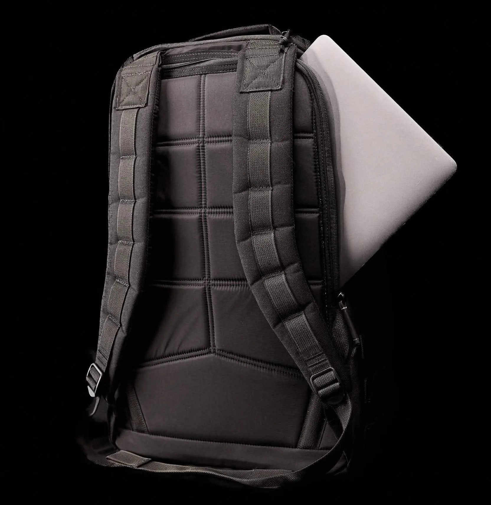 The back of the GoRuck GR1 is shown with a silver laptop peeking out from the side of the bag's rear laptop pocket. The bag is shown against a plain black background. 