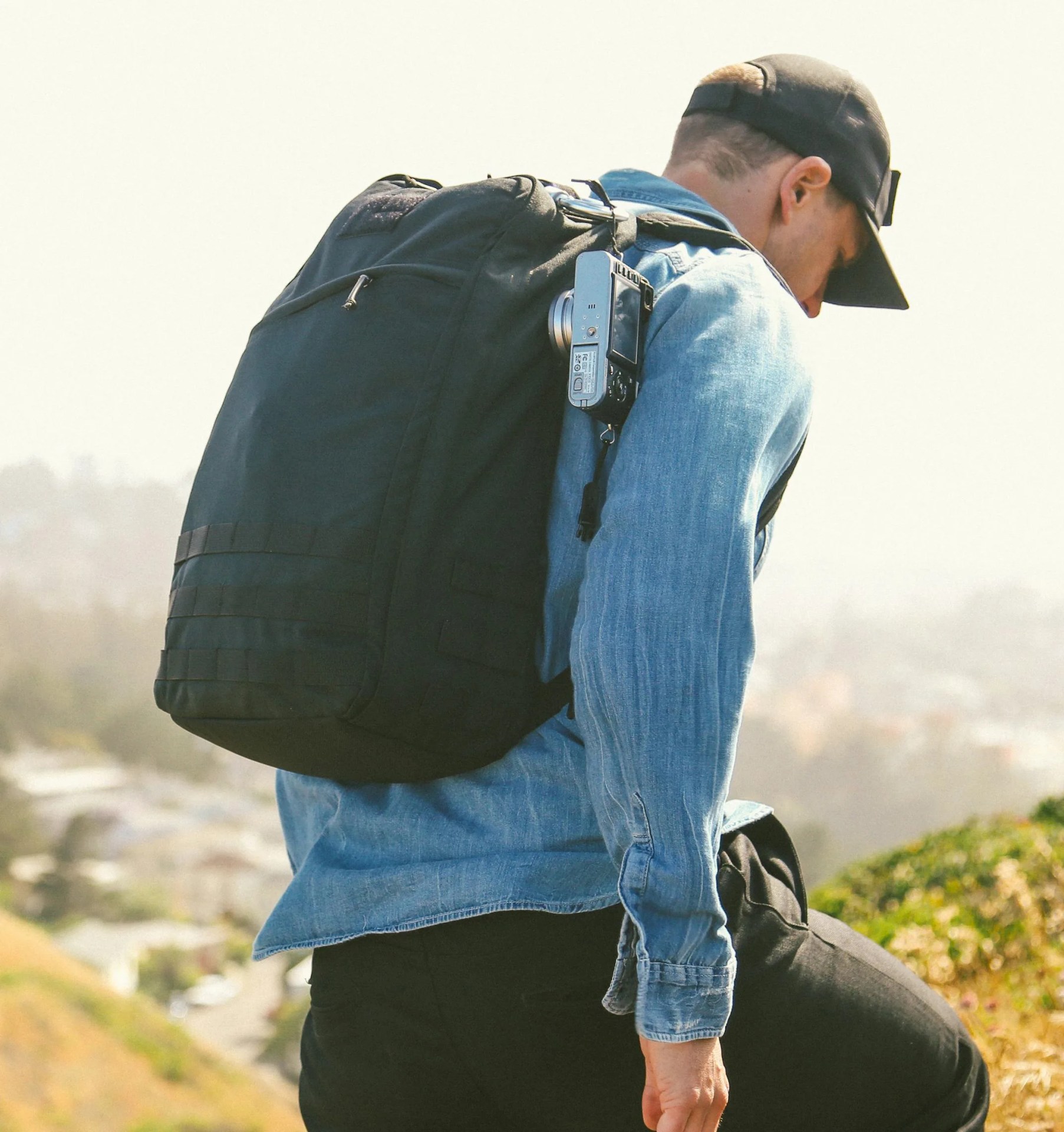 A man wearing a GoRuck GR1 is shown from behind. The man is wearing a denim shirt, black pants, and a black baseball hat. The GR1 is shown on his back. A handheld camera is also attached to the bag and hanging on the right- button-up shirt back in black. hand side. He appears to be walking on hilly terrain with blurred building appearing in distance. 