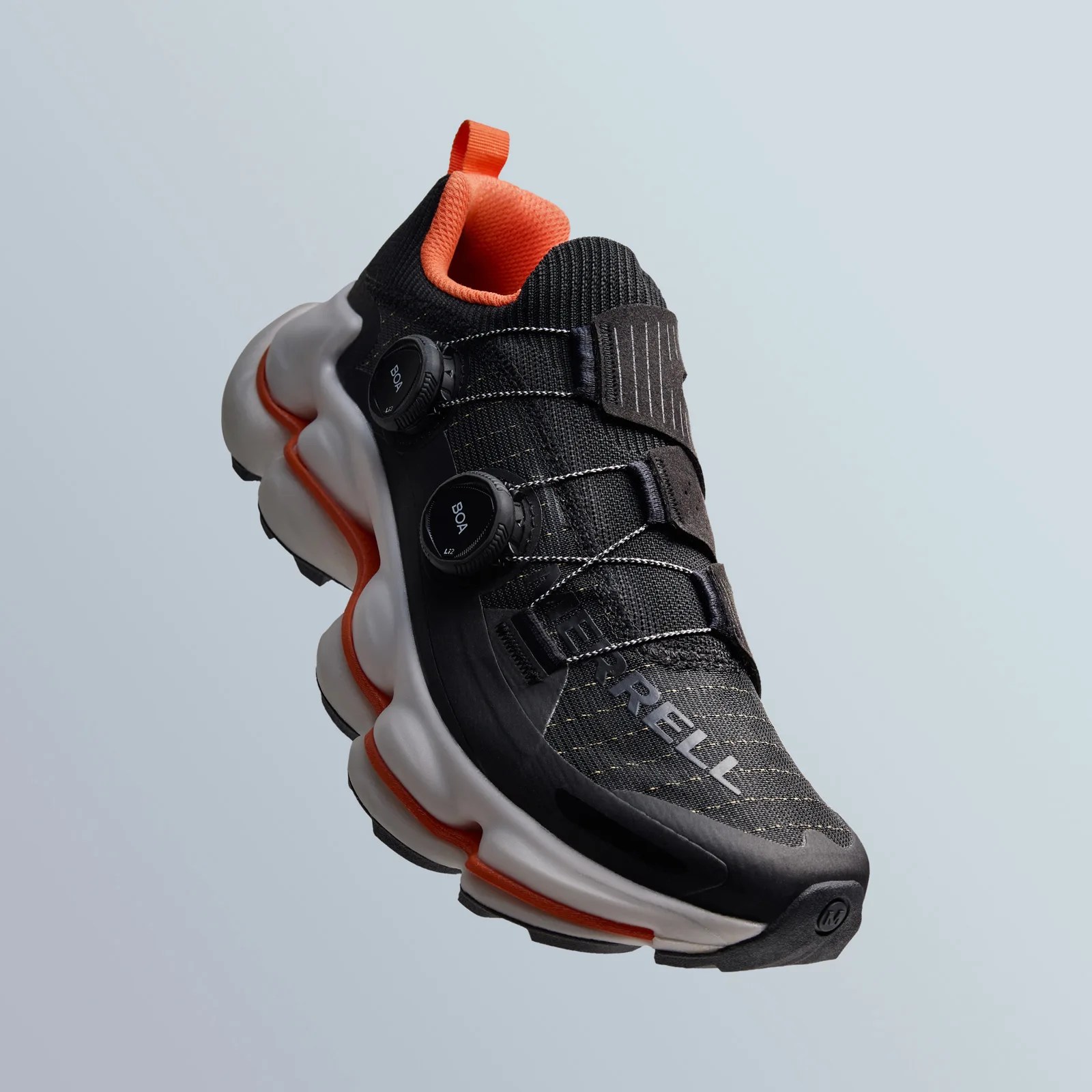 A shot of the Merrell SpeedArc hiking shoe. The shoe is black with orange details and a white sole. The shoe is angled in the shot to appear floating in mid air against a light grey background, with the toe pointing to the lower right corner of the image. 