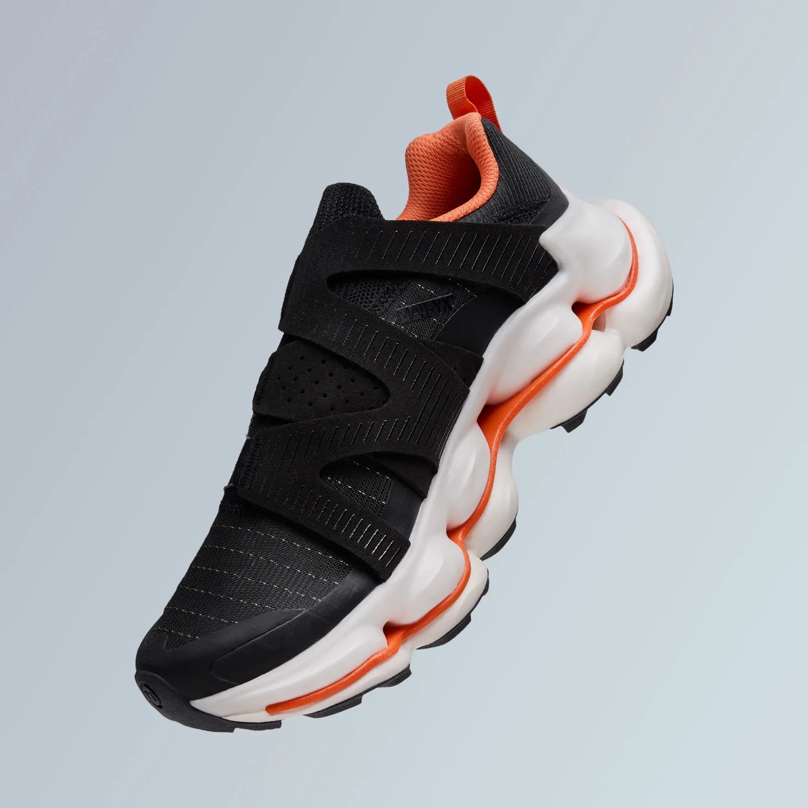 A shot of the Merrell SpeedArc hiking shoe. The shoe is black with orange details and a white sole. The shoe is angled in the shot to appear floating in mid air against a light grey background, with the toe pointing to the right right corner of the image, showcasing the outer upper and side of the sneaker.  
