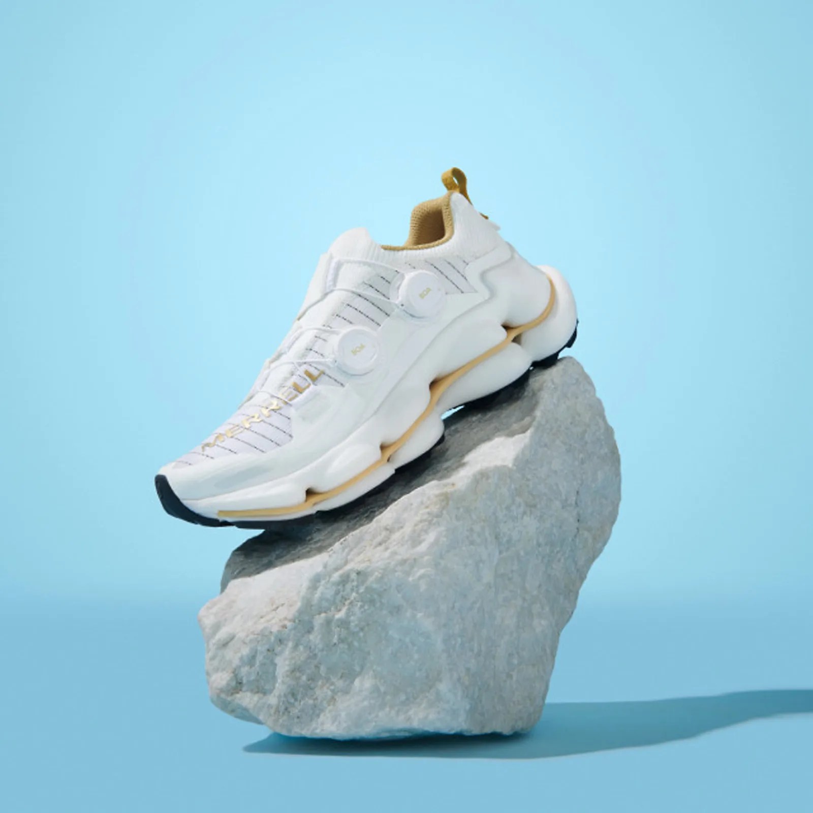 A single Merrell SpeedARC Surge BOA shoe shown positioned on a light grey rock with the shoe angled down so the toe is pointing to the bottom left corner of the image. The shoe itself is a white color with light gold / yellow detailing. The shoe and rock are shot on a light baby blue background. 