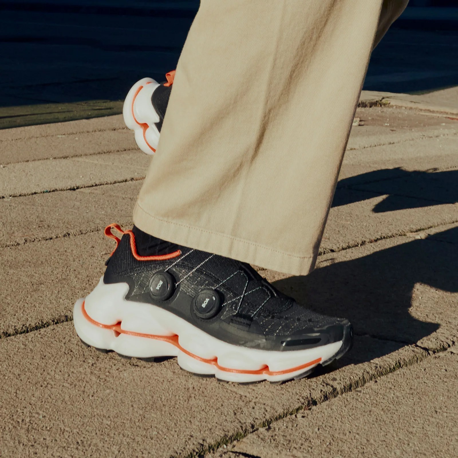 A pair of Merrell SpeedARC Surge BOA shoes worn on a model wearing khaki pants. The image shoes the shoes up close from the knee down. The right foot is shown touching the concrete stone ground from the side. The heel of the left sneaker is visible peaking behind the model's calf, lifted in mid-run / step. 