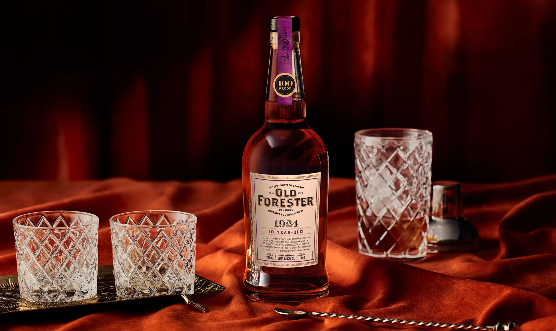 A bottle of Old Forester 1924 bourbon sitting on a velvety brown material flanked by crystal whiskey glasses and a metal cocktail stirrer. 