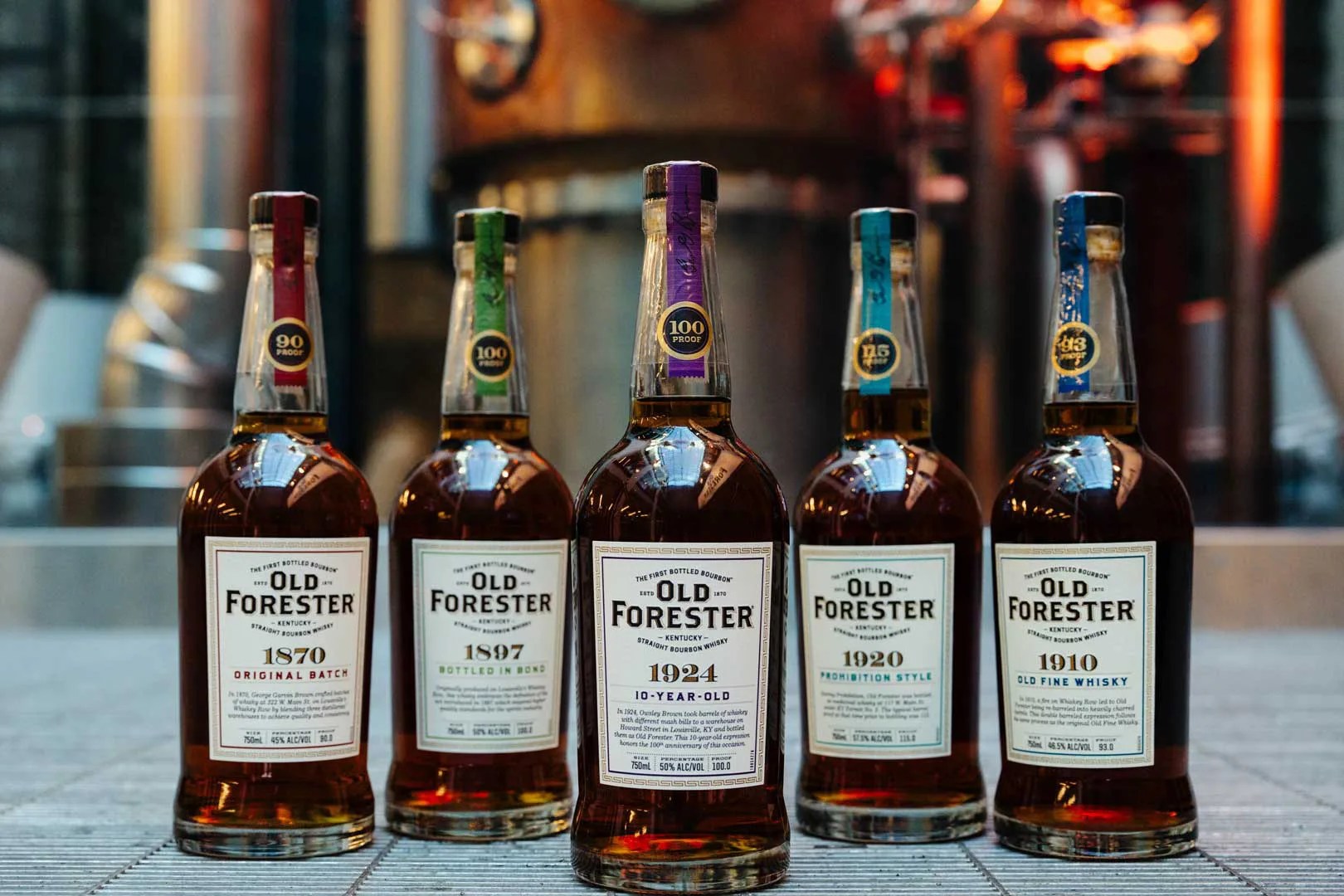 A collection of five Old Forester Whiskey Row bottles shown in front of distilling equipment. The 1924 bottle is positioned in front of the other four bottles.