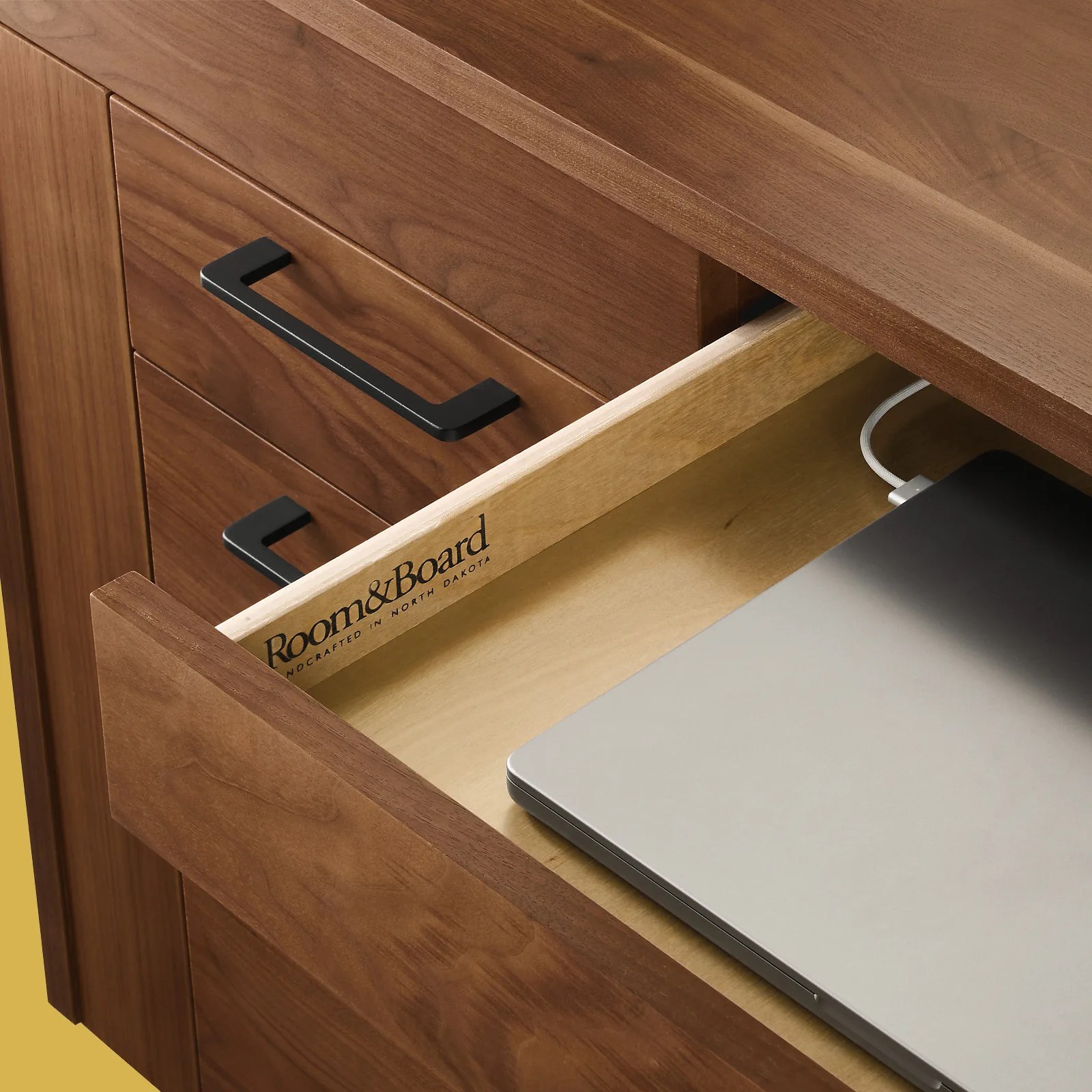 A closeup view of the laptop drawer located in the center of Room&Board's Lincoln standing desk. The drawer is shown partially open to reveal a laptop charging in the drawer. 