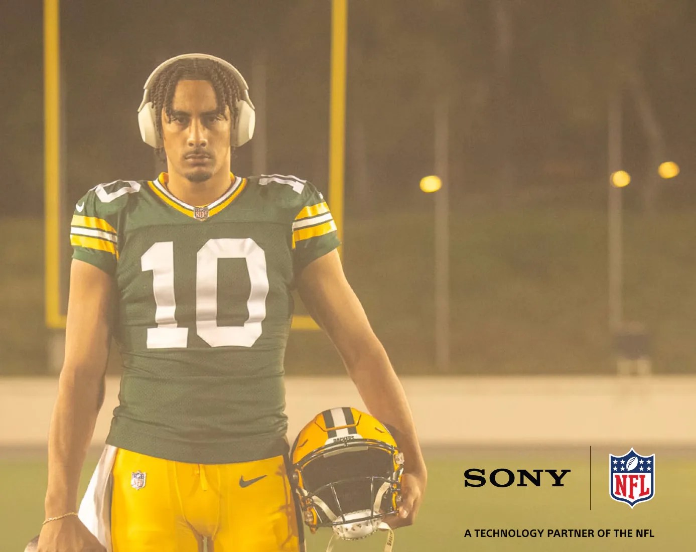 An image of an NFL player in a Green Bay Packer's uniform holding his helmet on the right side of his body. He is standing on football field with a set of uprights behind him. A Sony and NFL logo appearing in the right hand corner of the image. 