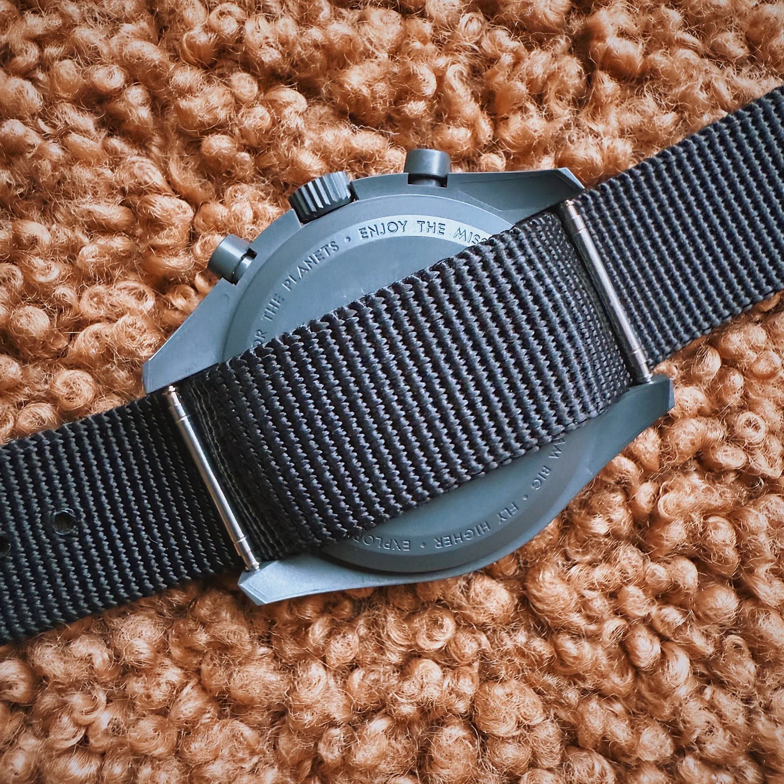The back of a Swatch MoonSwatch Mission to Mercury shown to reveal a black Nato strap threaded through two spring bars. The reverses watch is resting against a fuzzy brown material.