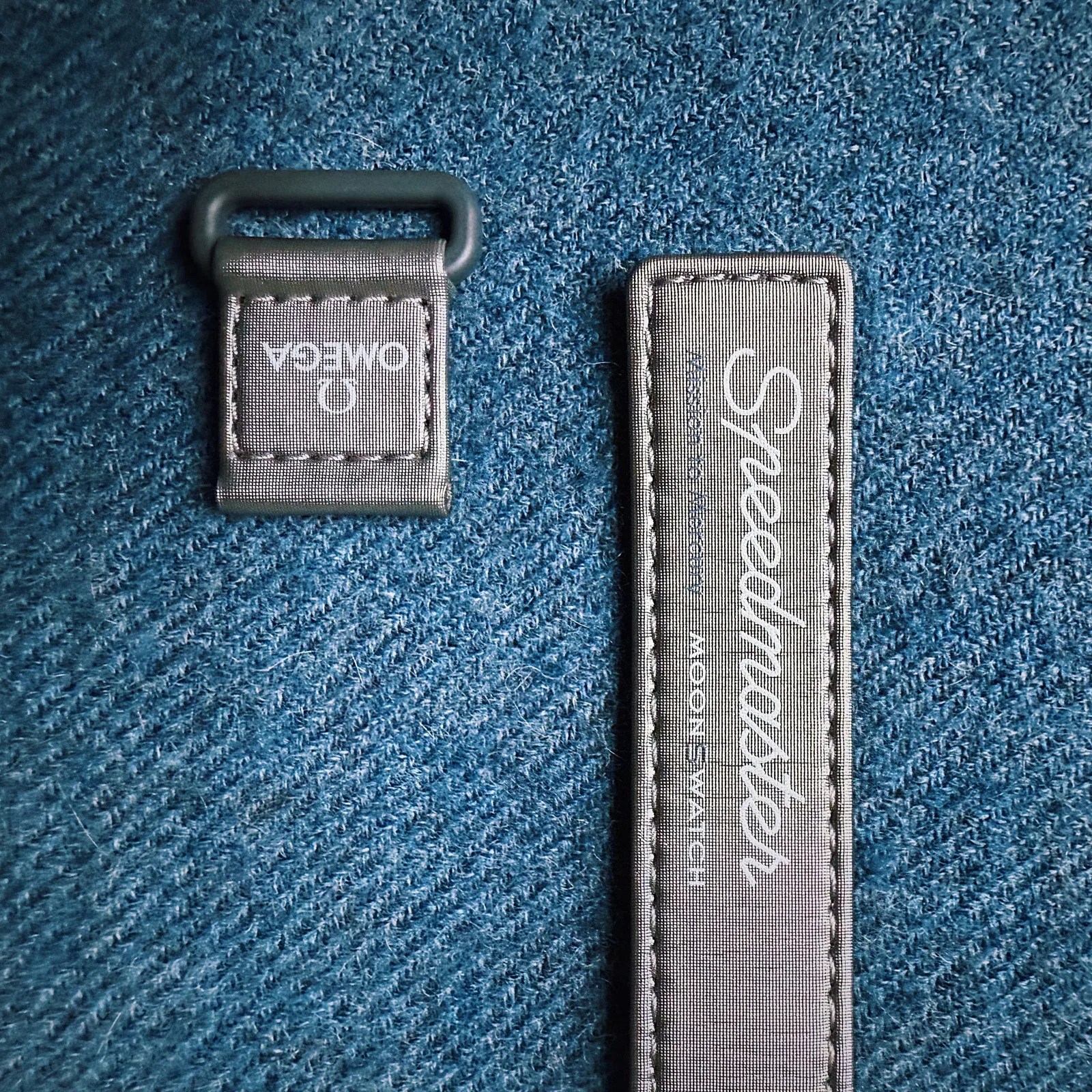 The two pieces of the Swatch MoonSwatch strap shown laying against a light blue fabric background. The strap matches the Mission to Mercury MoonSwatch and is a light silver in color. 