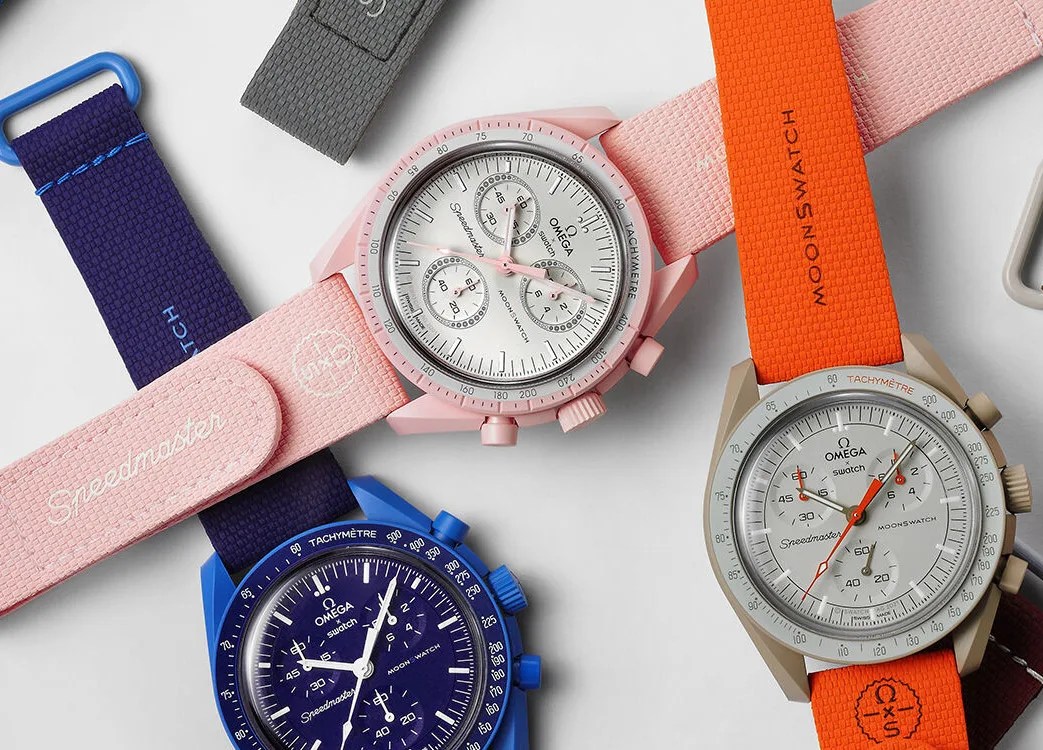 Three Swatch MoonSwatches in various bright colors shown laying in a pile on a light grey surface. Each bold colored watch is paired with a brightly colored matching rubber strap in colors of dark blue, light pink and blaze orange. 