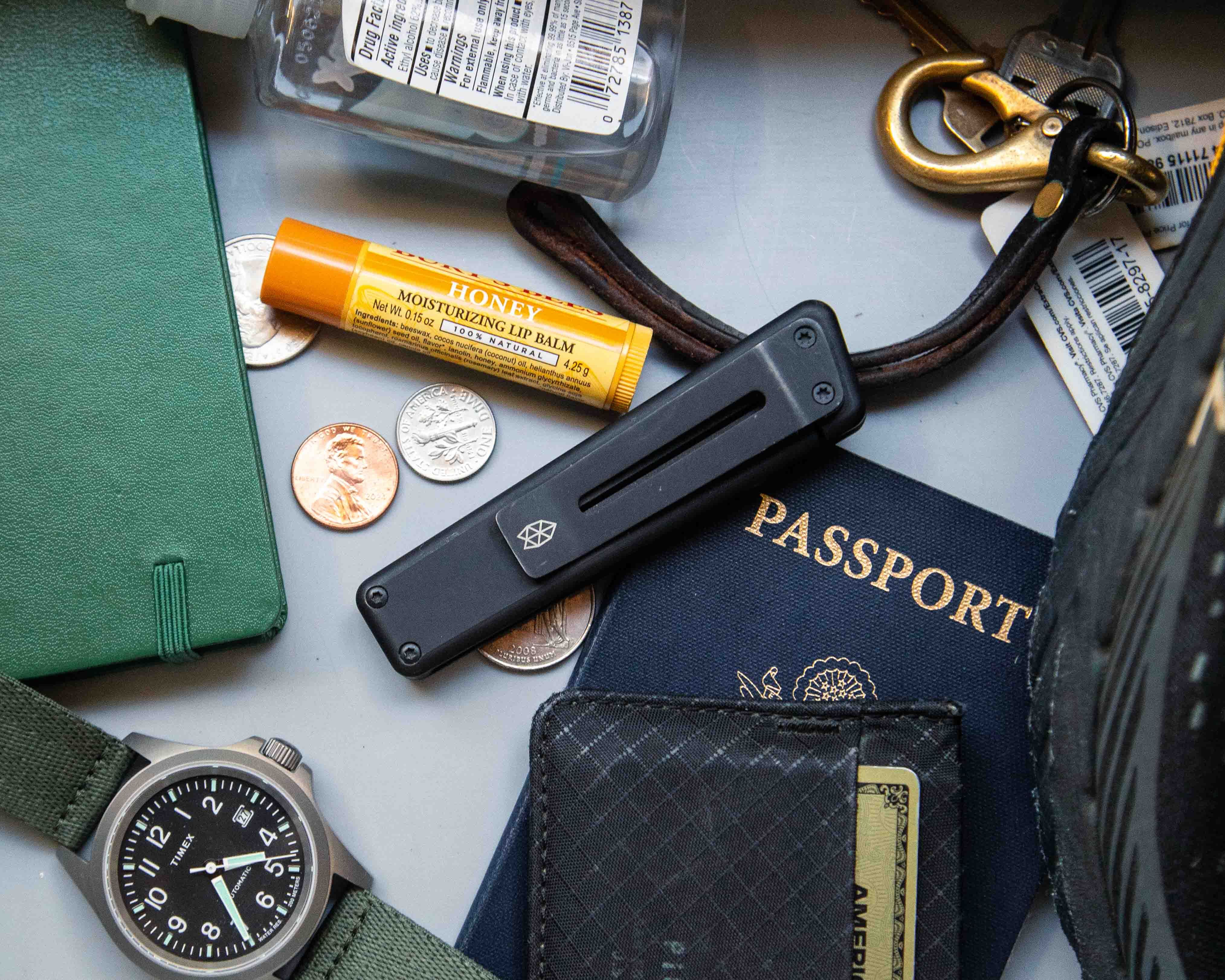 James Brand Chapter knife surrounded by lip balm, coins, keychain, hand sanitizer, Pioneer Carry wallet, passport and notebook on gray background