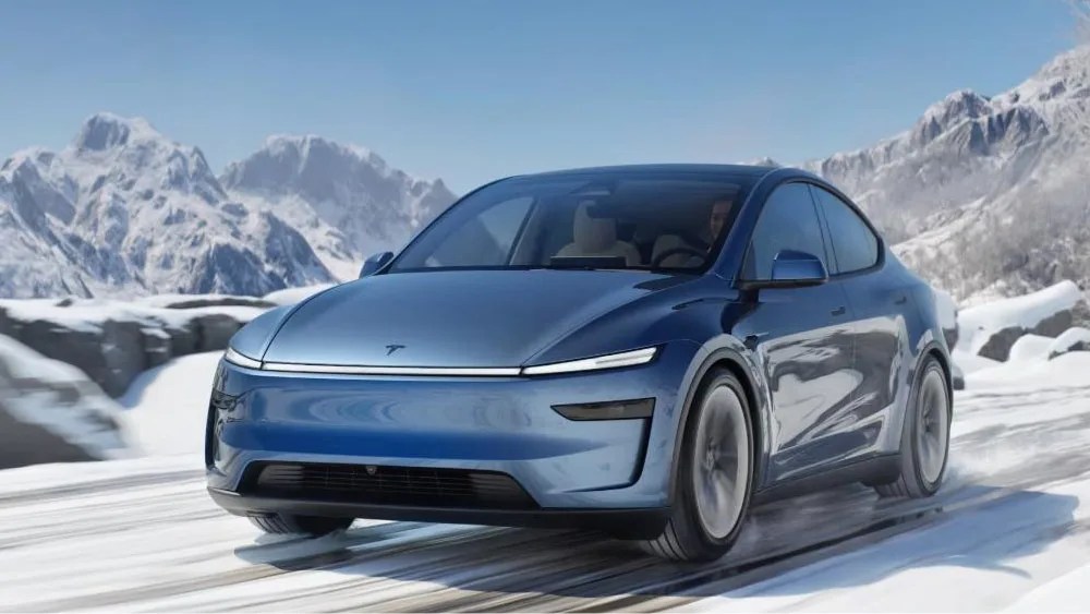 A 2025 Tesla Model Y shown in a new Glacier Blue color way shown driving on a snowy road in the daytime with the front headlights lit. 