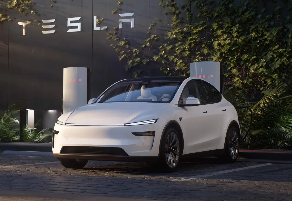 A white updated 2025 Tesla Model Y shown parked and charging in a parking lot next to a large gray building with Tesla written on the wall. 