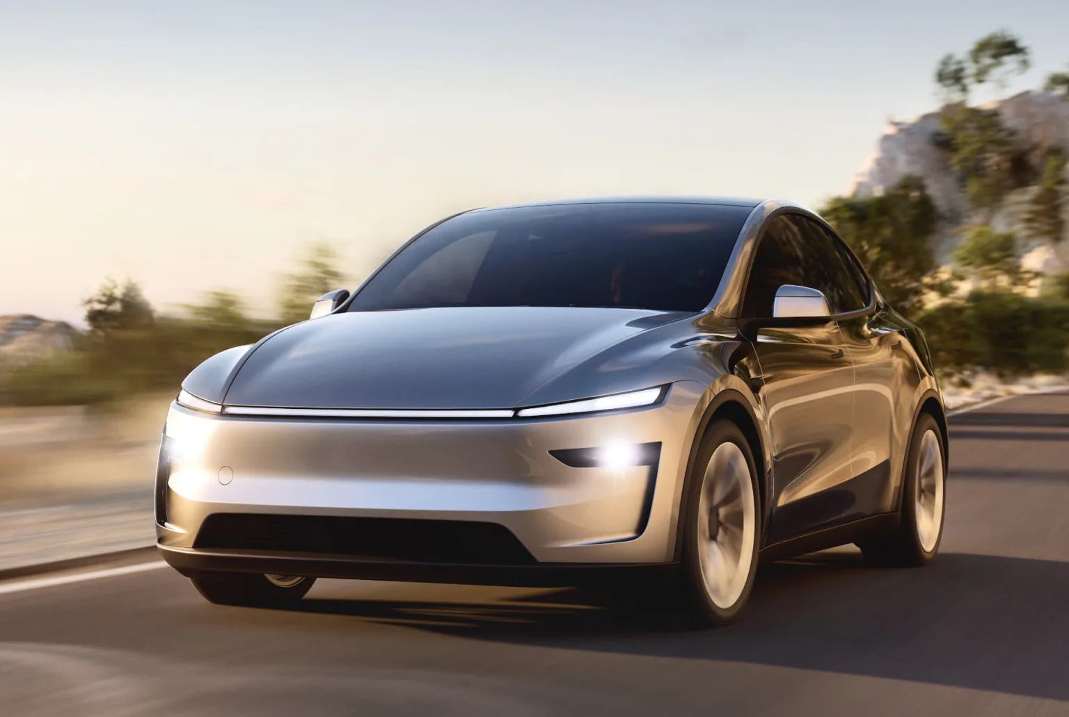 New Tesla Model Y for 2025 in silver shown driving along a road with the headlights on. Palm trees and blue sky appear blurred in the photo background.