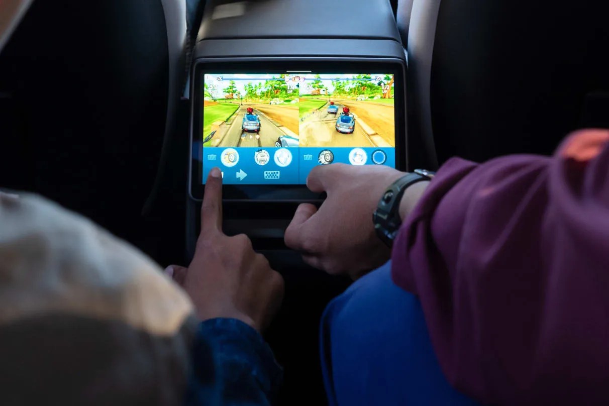 An image of the rear screen in the new Tesla Model y 2025. Two sets of arms and hands are shown interacting with the small lit display playing a racing game. 