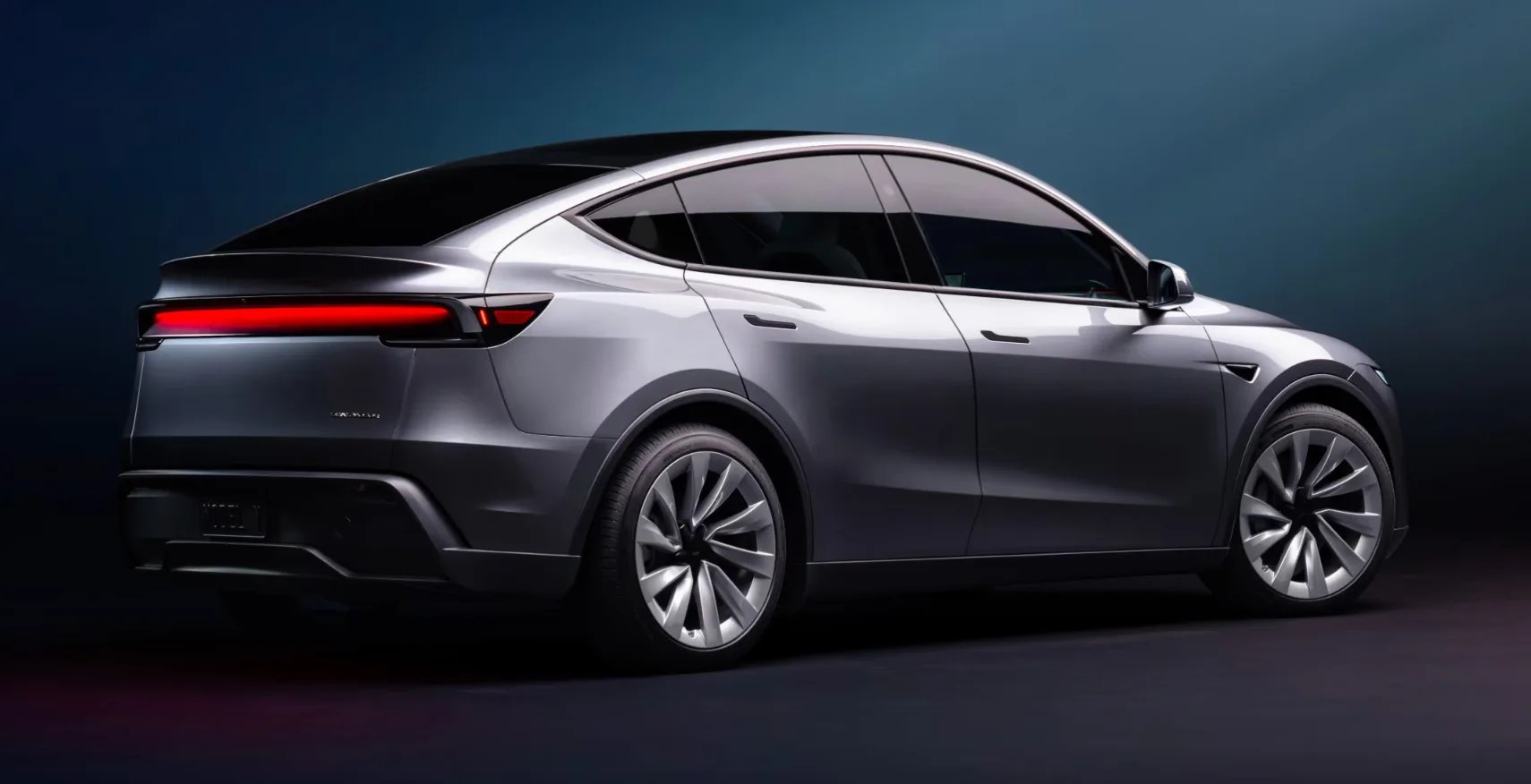 Tesla Model y 2025 edition shown sitting on a grey surface against a dark blue background. The car is dark grey and positioned so that the rear right corner is closest, with the side of the car extending out and to the right of the picture. 