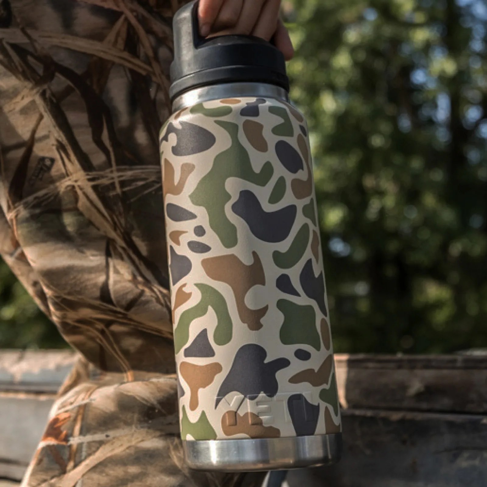 A Yeti Rambler water bottle being held in a hand hanging next to a person's leg. The person is wearing camouflage in the woods. The Yeti Bottle shown is in a Tan Camo color.