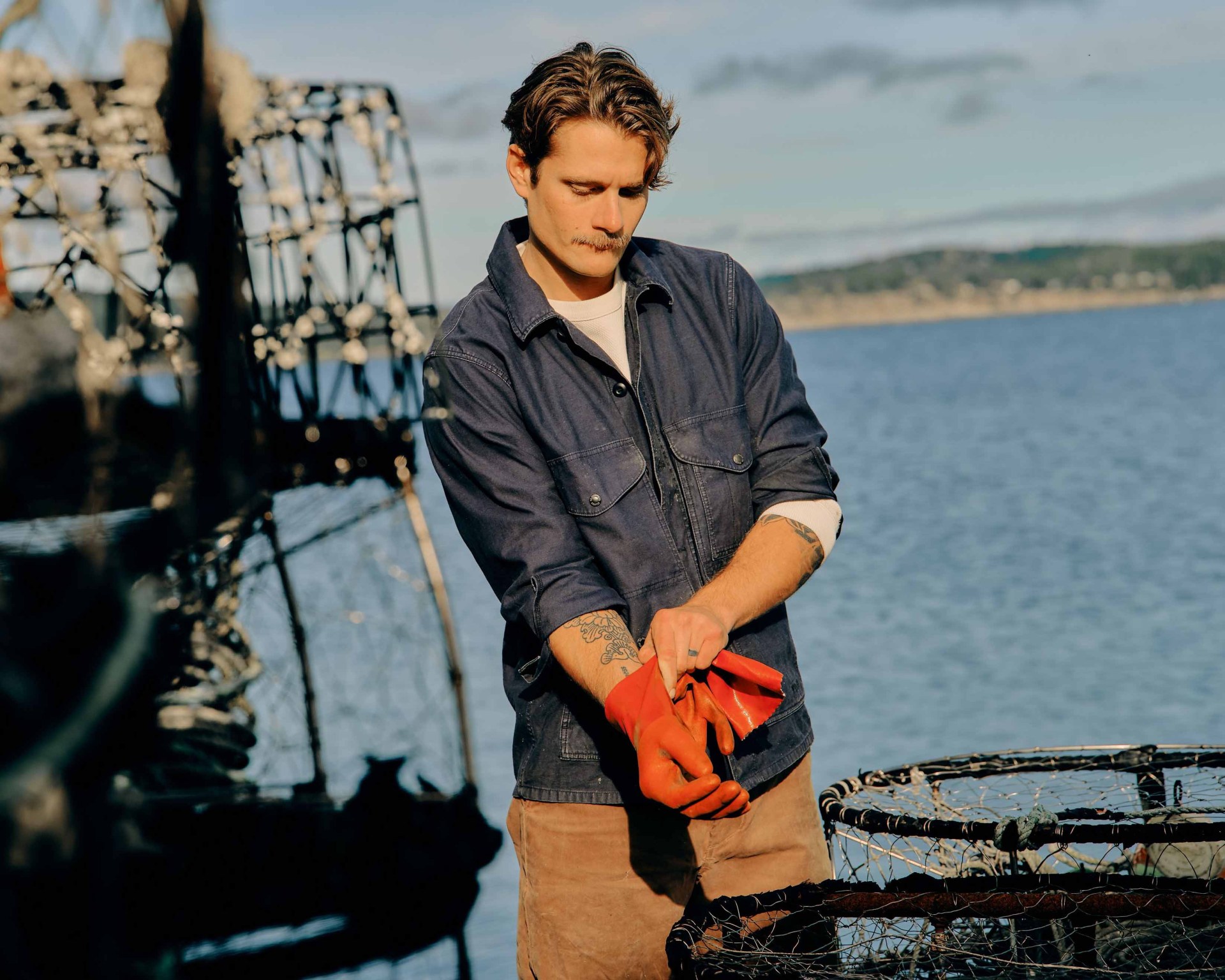 a man on a dock wearing a blue shirt, putting on orange gloves