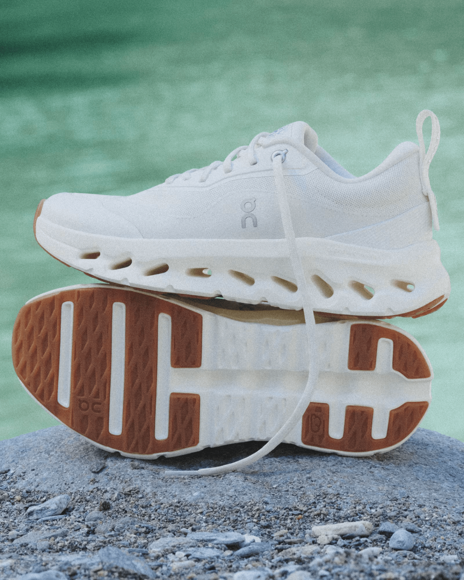 A pair of 2025 On Cloudtilt Loewe 2 running shoes in all white. The shoes are resting on a rocky surface with what looks to be a body of teal green water blurred in the background. 