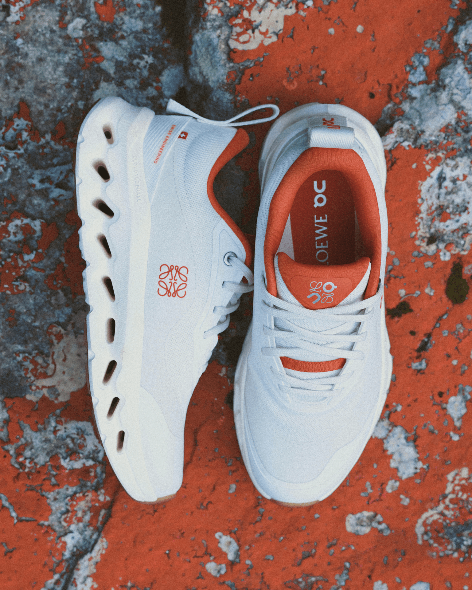 A pair of 2025 On Cloudtilt Loewe 2 running shoes in a burnt / reddish orange shown empty side by side with one sneaker laying on its side to show the exterior panel and Loewe logo. The shoes are resting on a rocky surface covered in a matching orange paint / moss. 