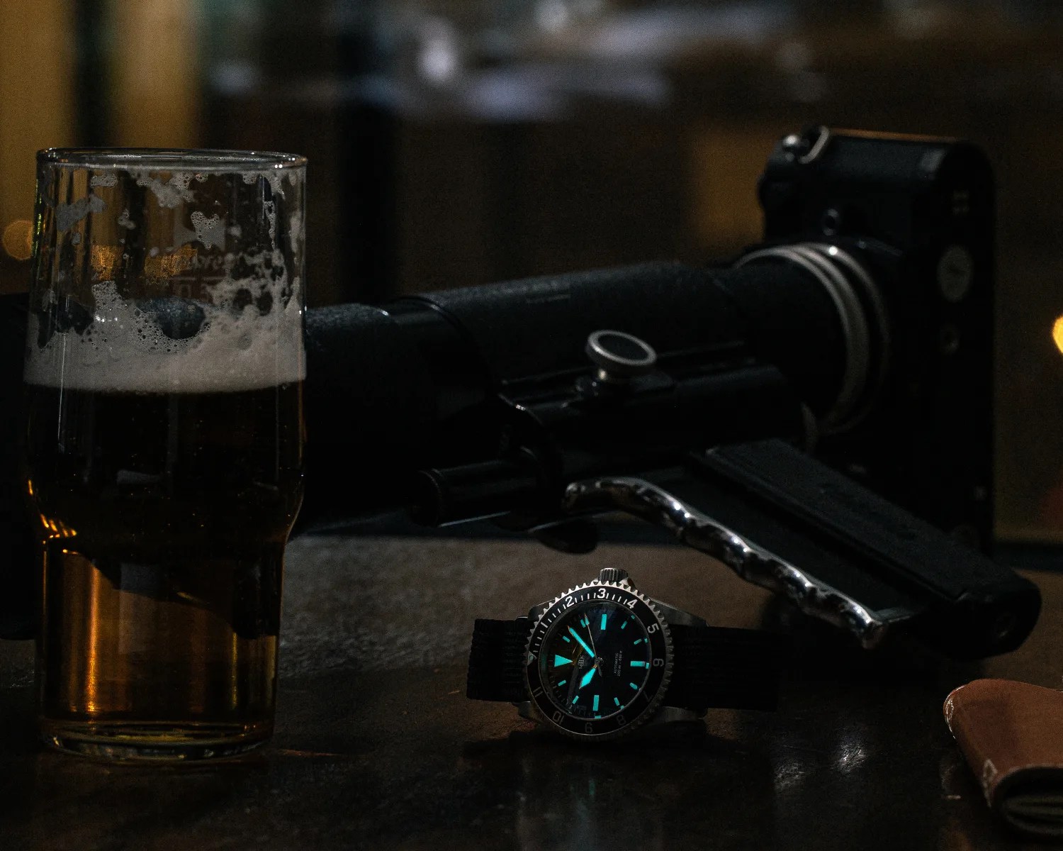a Mk II dive watch glows in the dark on a bar next to a camera and a glass of beer