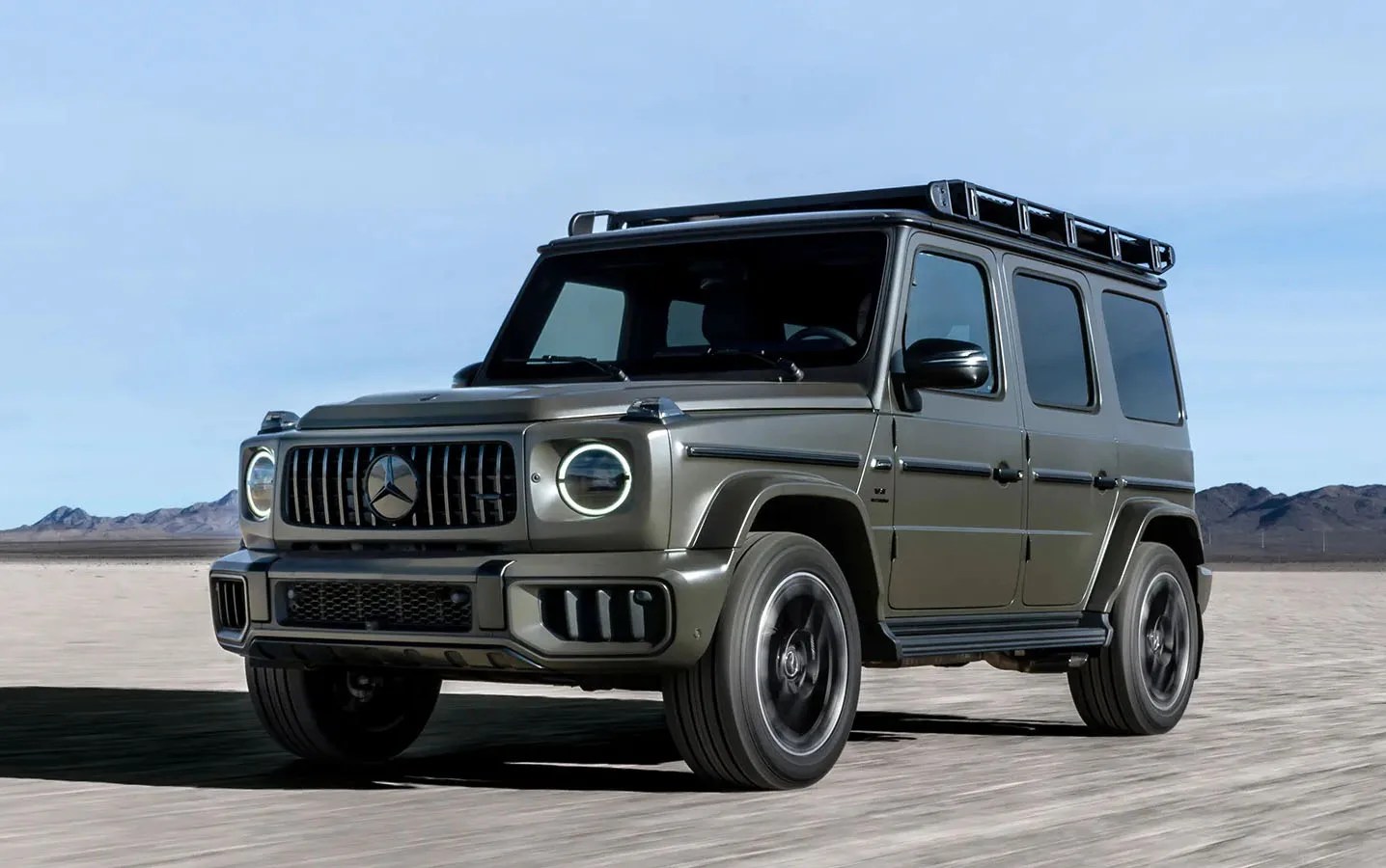 The 2025 Mercedes-Benz G-Wagen in a dark grey color shown driving on a flat desert landscape