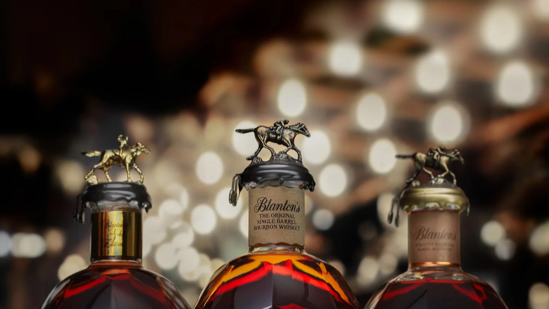 Three tops of different Blanton's Bourbon bottles shown from the neck up against a blurred background of twinkly lights
