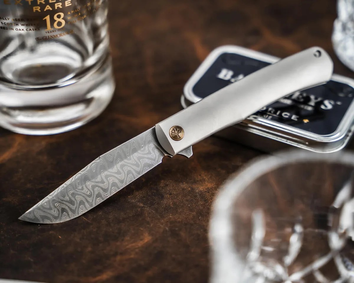 Böker Damast Annual Knife 2025 on leather table with tin, crystal glasses and scotch bottle