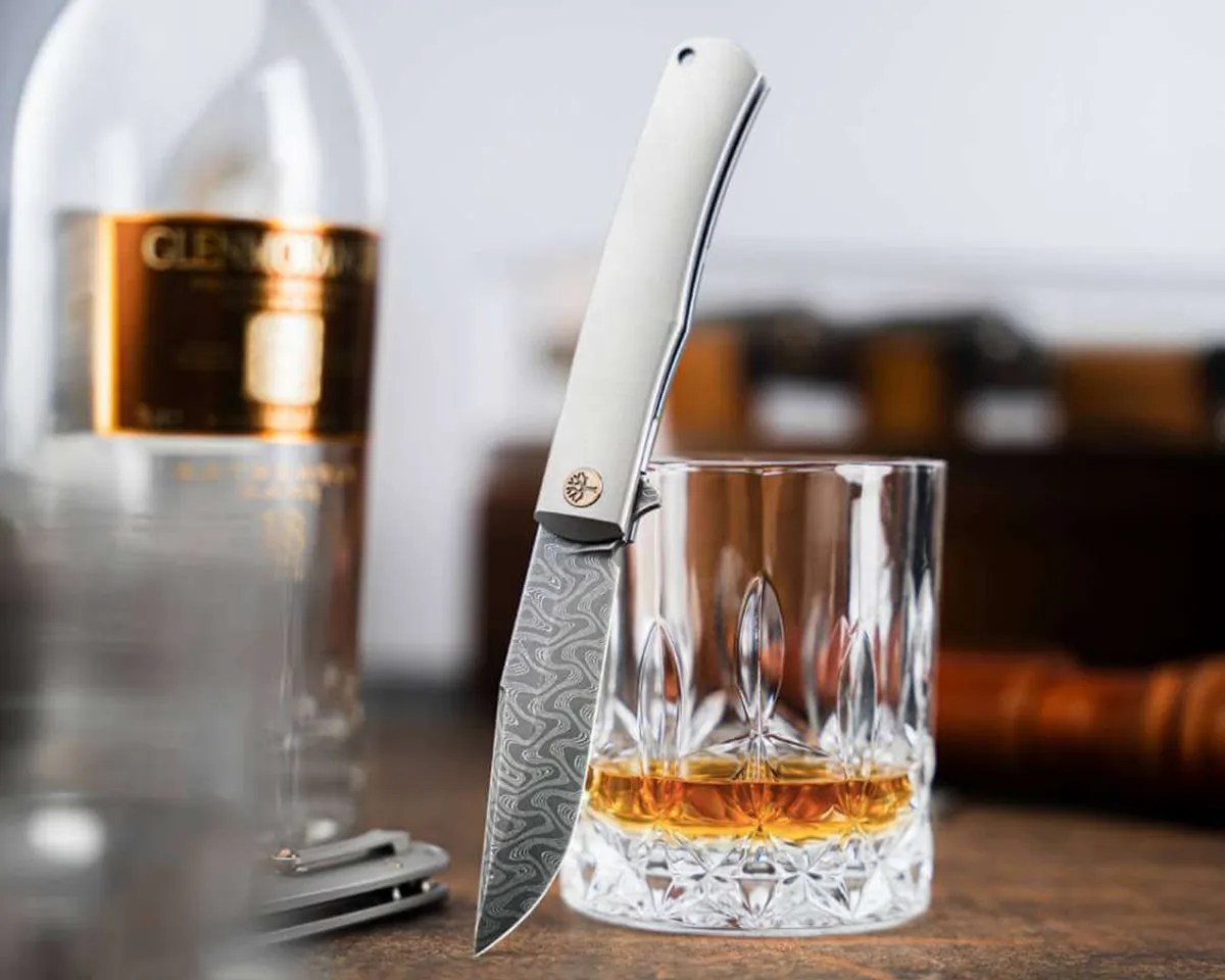 Böker Damast Annual Knife 2025 on leather table with tin, crystal glasses and scotch bottle