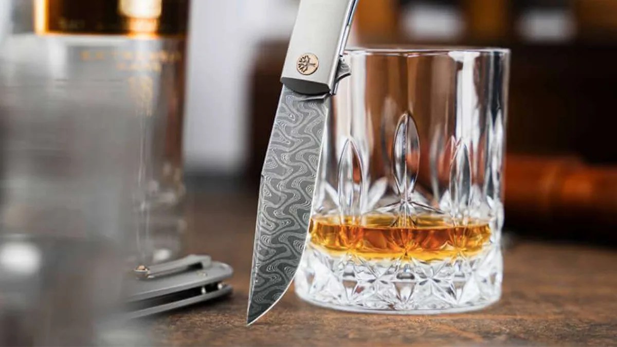 Closeup of Böker Damast Annual Knife 2025 on leather table with tin, crystal glasses and scotch bottle