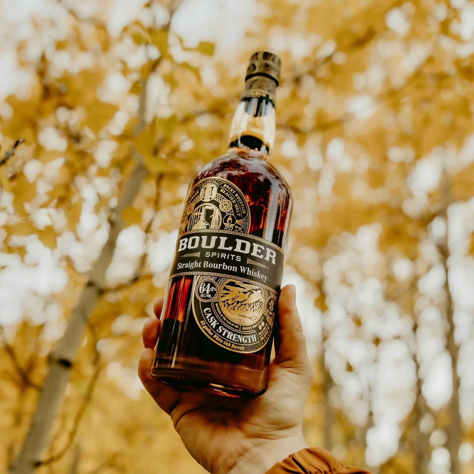 A bottle of Boulder Spirits Cask Strength Straight Bourbon Whiskey is held in hand up against a blurredbackground of a forest of trees with yellow leaves. 