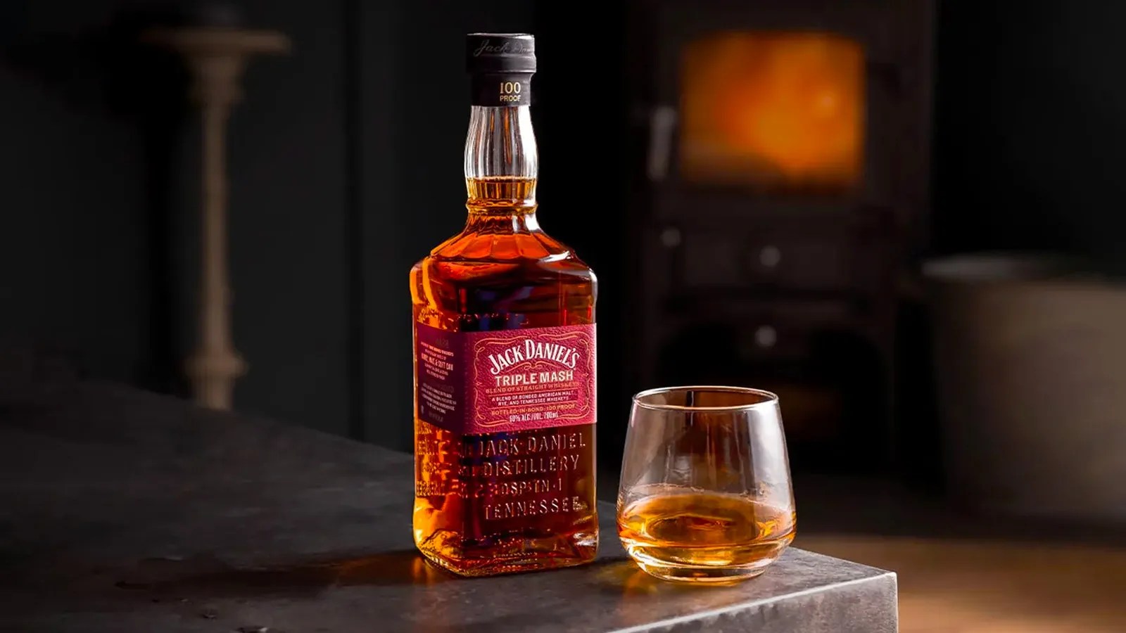 A bottle of Jack Daniel's Triple Mash Blended Straight Whiskey sitting on the corner of a metal table next to a rocks glass filled with whiskey. The dark room is blurred behind the bottle and shows vague details of a pillar and container.