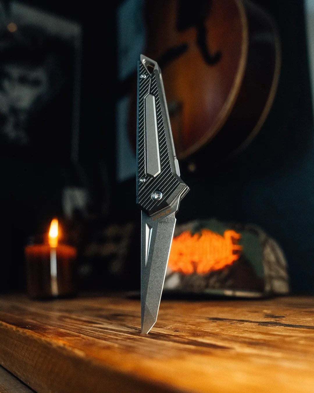 Pichi Design Kameta knife stuck in wood table in front of camo hat, candle and guitar