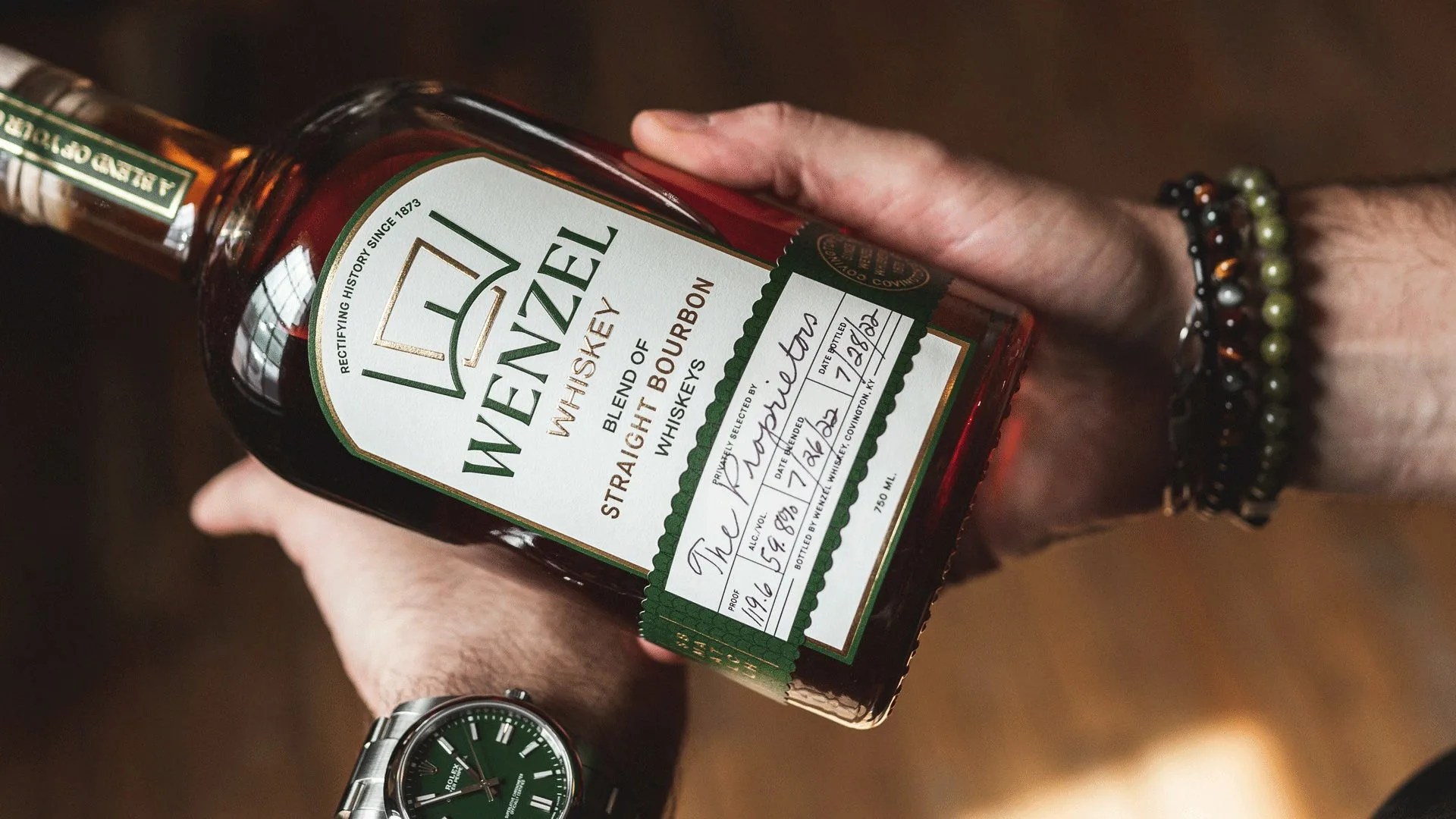A bottle of Wenzel Whiskey being held in the hands of a man wearing a watch and bracelets. The bottle label is white with green labeling with gold labeling.
