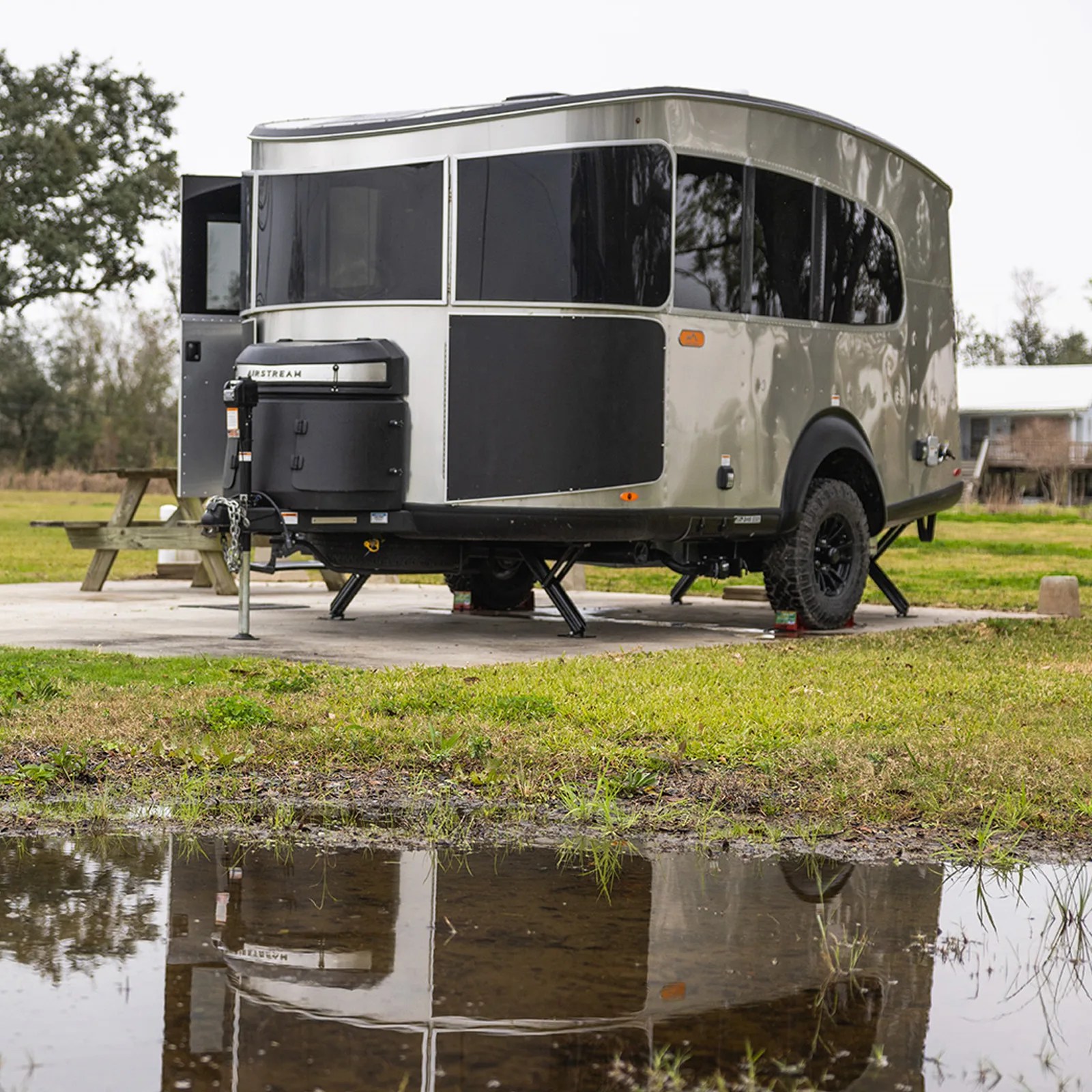 The Airstream Basecamp Xe adventure trailer is shown parked on a concrete pad next to a picnic table in front of a small pond, which reflects the trailer. 