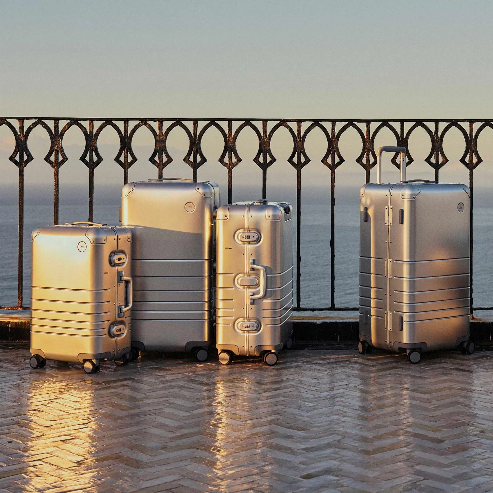Four Monos Aluminum hard-sided suitcases of various sizes standing up and clustered together against a wrought iron railing on a balcony with the ocean in the background. Warm sunlight is shining on the suitcases from the left hand side of the image. 