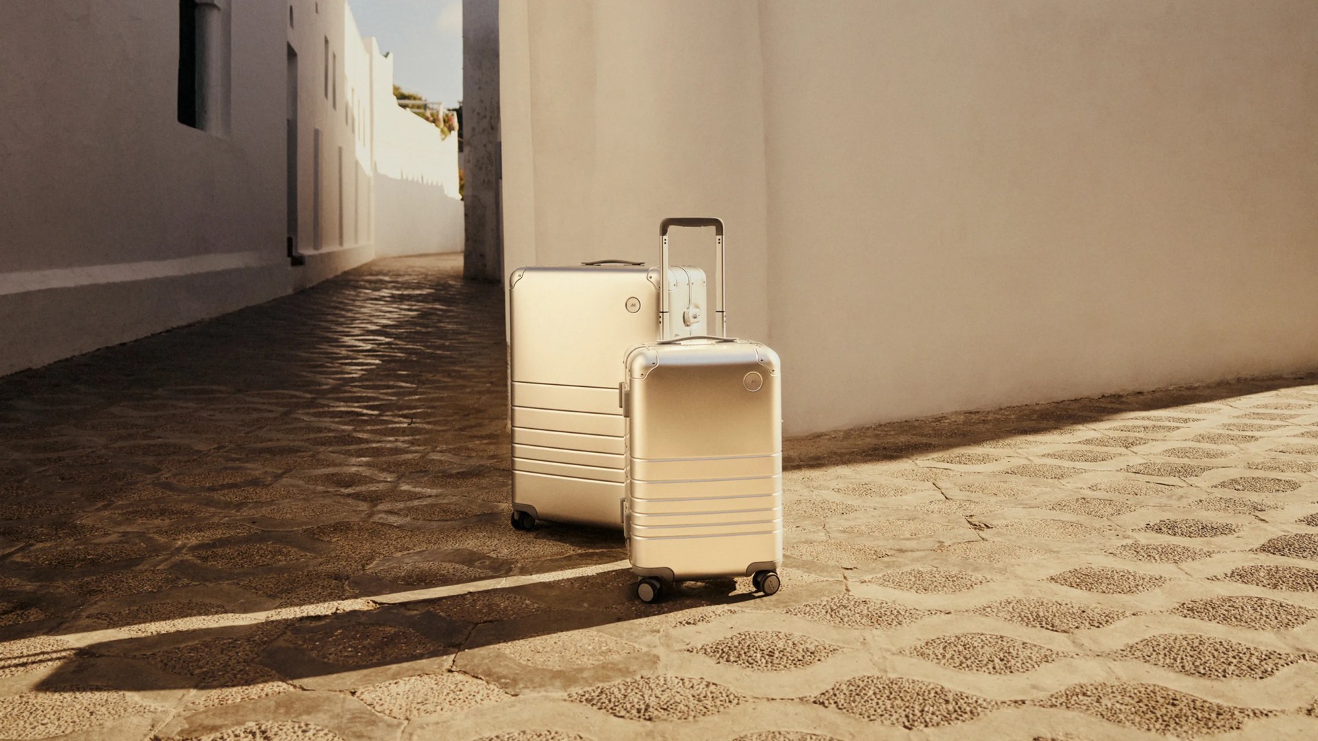 Two Mono all aluminum hard-sided suitcases shown in an empty street paved in stone. Both bags are lit by warm sunlight from the right hand side of the image casting long shadows.