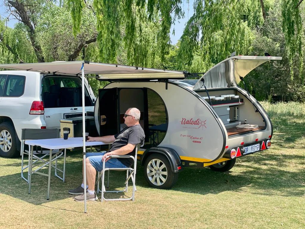 A Naledi Camper shown parked with the rear kitchen hatch open and an integrated awning extended. An older man with white hair is shown sitting beneath the awning in a folding chair next to a camp stable. 