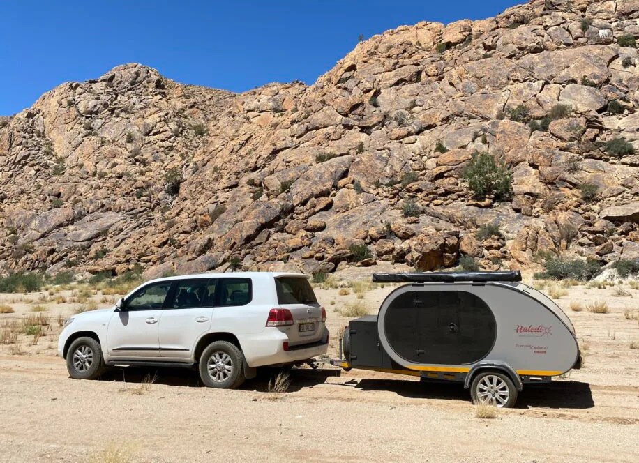 A Naledi Campers being towed by a white Toyota SUV across a sandy landscape with a rocky hill in the background.