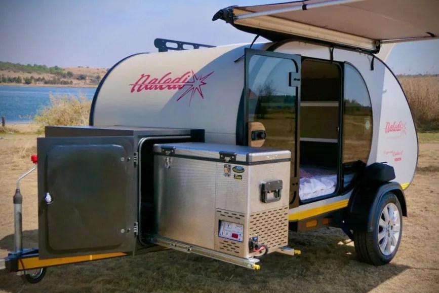 A Naledia Camper shown parked with the door open and integrated roof awning pulled out. The camper's combination fridge / freezer is shown pulled out on its storage rails for access. 
