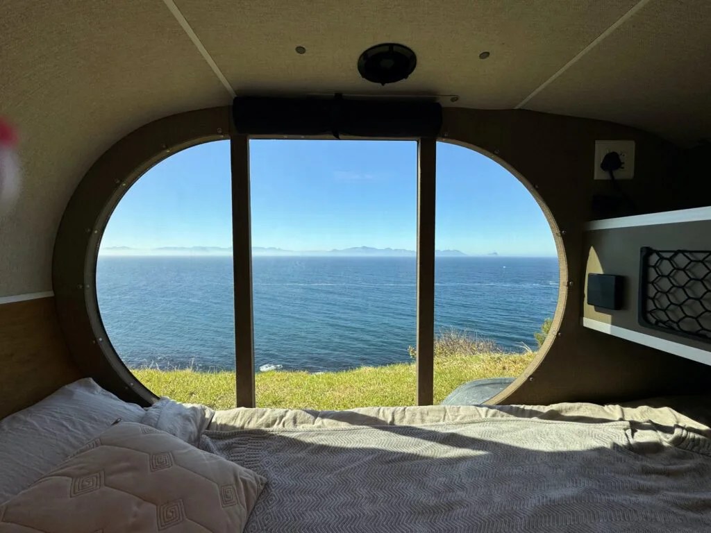 A interior view of the Naledi camping view highlighting the adventure trailer's sizeable tinted window, which  offers a wide view of the ocean and green grass around the trailer