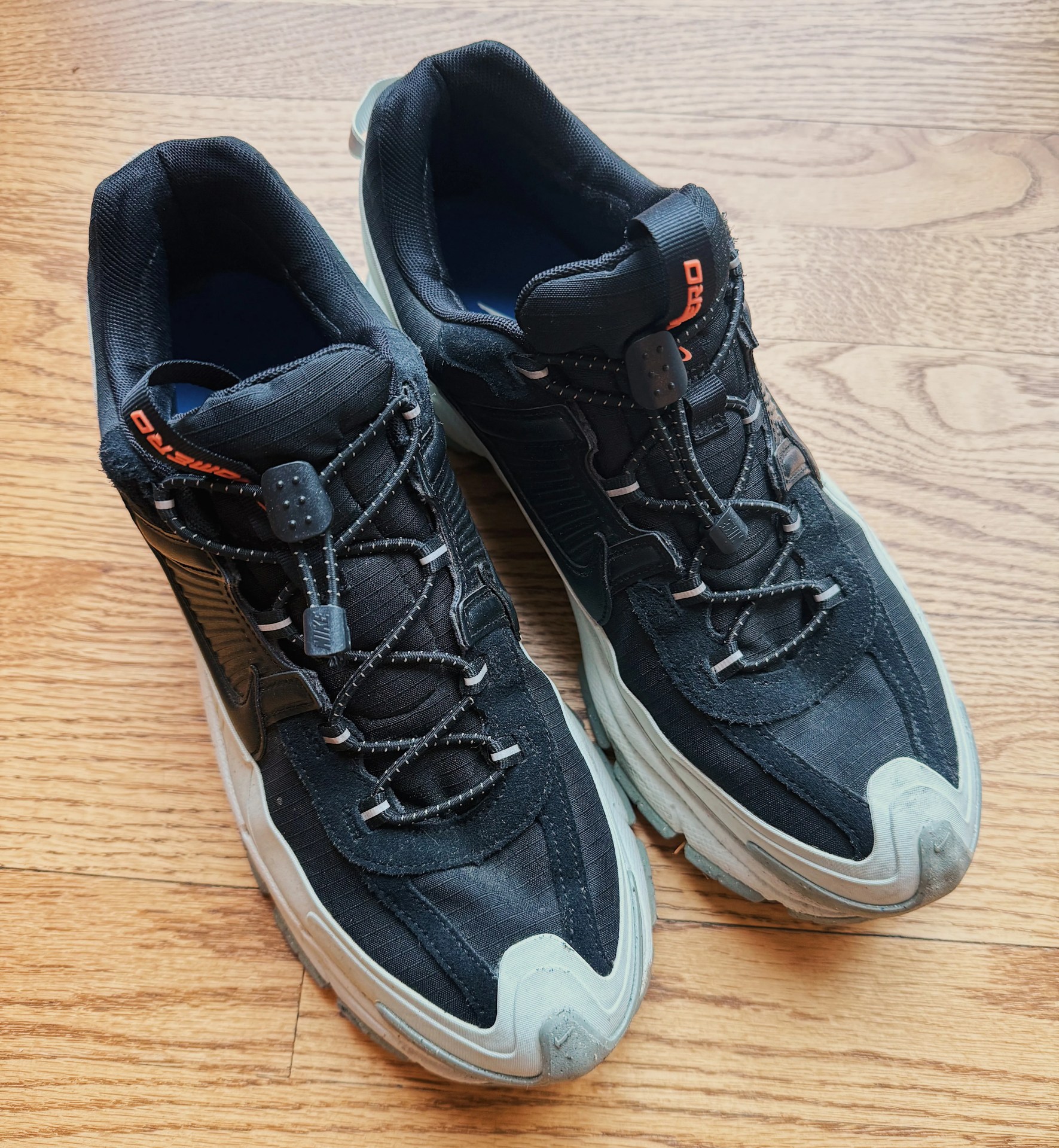 A pair of Nike Zoom Vomero Roam sneakers in black sitting side by side on a light wood floor. 
