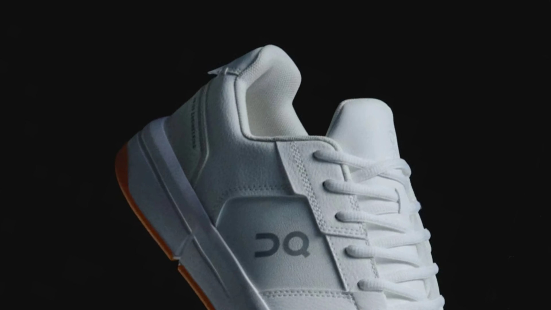 A close up of the heel of the ON THE ROGER Clubhouse revised sneaker. The sneaker is white with a tan colored gum sole and shown against an all-black background.