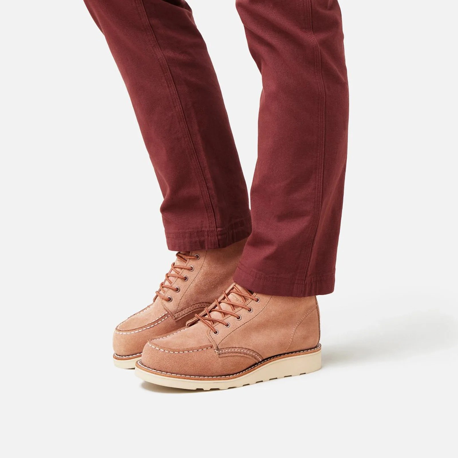 A pair of Red Wing Heritage Classic Moc boots in a new Dusty Rose color, which looks like a suede-like mix of pink and red, being worn by a female model wearing dark marroon pants. The pants and boots are shown from right below the knee down. 