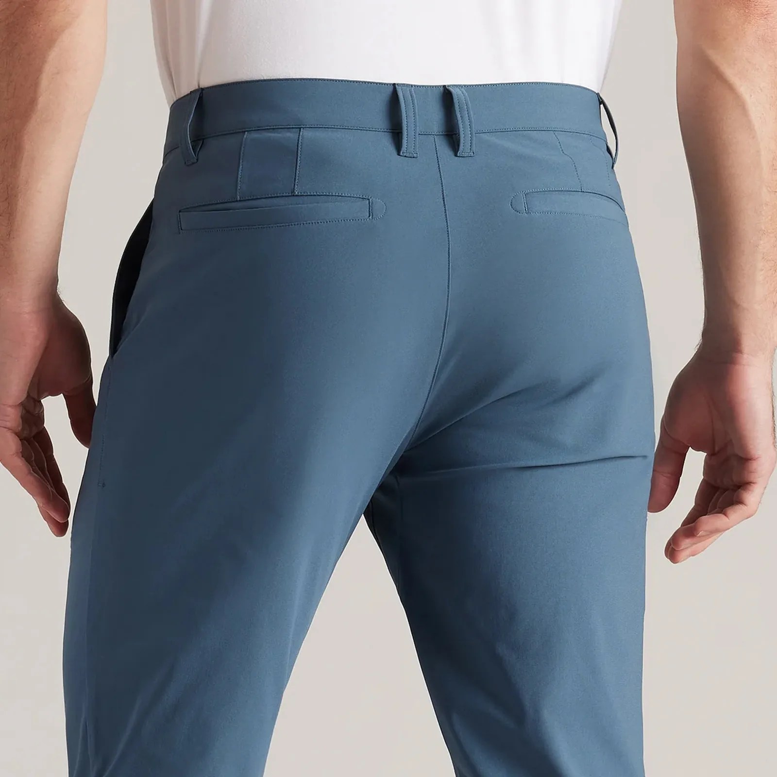 A close up of the rear butt area of the Rhone Commuter Pant in a light blue. The pants are being worn by a light-skinned model shown from the waist to knee area facing backward. The models arms are shown hanging down. 
