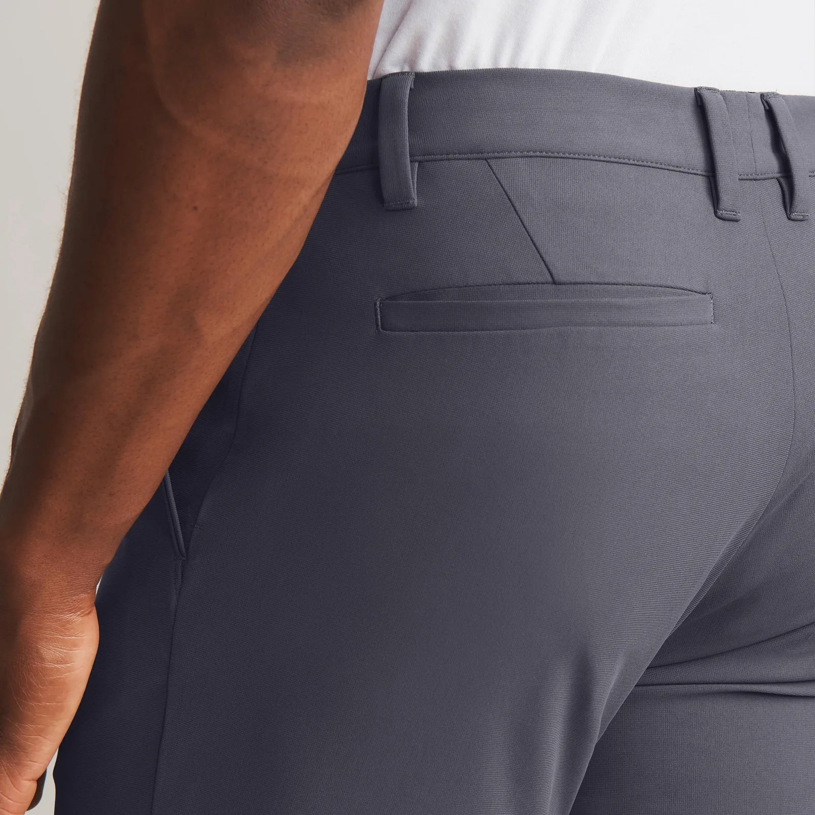 A close-up view of the rear left pocket of the Rhone Commuter Pro dress pants in light grey being worn by a dark-skinned male model whose lower fore arm is visible and resting against the pant pocket. 