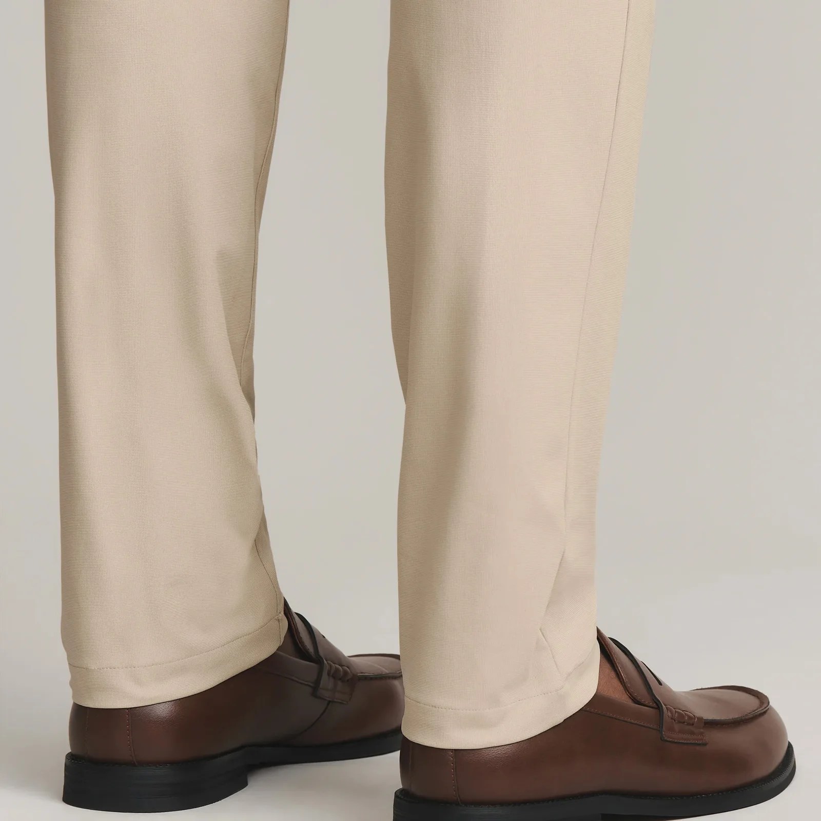 A view of the ankle and lower leg area of the Rhone Commuter Dress Pant Pro in light sandy / khaki. They are shown on the legs of a model from below the knee down. The model's feet are facing backward to the right of the image. The model is wearing brown colored loafers with no socks. 